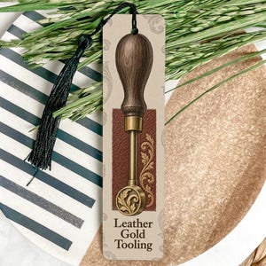May include: A rectangular bookmark featuring a graphic illustration of a leather gold tooling tool with a dark wooden handle and a brass tip. The bookmark has a cream background with subtle scrollwork and a black tassel attached at the top.