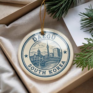 May include: A round, cream-colored ornament with a blue illustration of the city of Daegu, South Korea. The ornament has the text "Daegu South Korea" in a circular design. A gold string is attached for hanging.