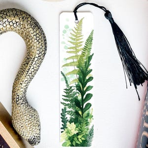 May include: A white bookmark with a black tassel features a watercolor illustration of green ferns and leaves.