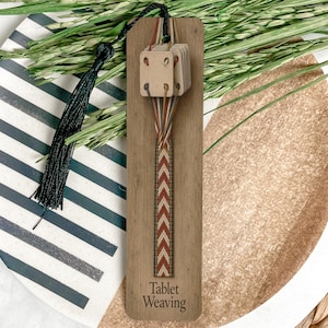 May include: A wooden tablet weaving tool featuring a rectangular base with the text Tablet Weaving engraved at the bottom. It includes a set of small square cards with four holes each, threaded with brown, white, and orange yarn in a chevron pattern.