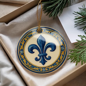 May include: Round ceramic ornament featuring a blue fleur-de-lis design on a cream background, framed by a blue and gold decorative border. The ornament hangs from a gold string and is presented in a gift box.