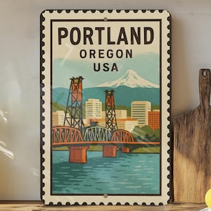 May include: A vintage-style metal sign featuring a scenic illustration of Portland, Oregon, USA, with the city's name and a bridge over water. The sign has a postage stamp design with a mountain in the background.