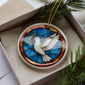 May include: Round ceramic ornament featuring a white dove holding an olive branch, set against a stained-glass style background of blue, orange, and red. The ornament is in a gift box with a gold string for hanging.
