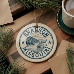 May include: A round, cream-colored ceramic ornament with a blue illustration of a riverboat on a lake, mountains, and the text "BRANSON MISSOURI". The ornament has a gold string for hanging and is surrounded by pine needles.