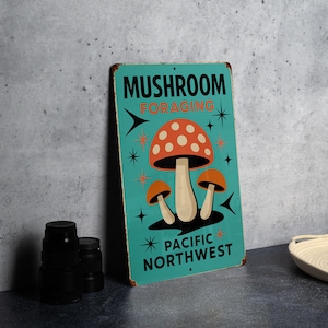 May include: A rectangular metal sign with a teal background features a retro illustration of three mushrooms. The sign reads "MUSHROOM FORAGING" and "PACIFIC NORTHWEST" in black text. The mushrooms have orange caps with white spots.