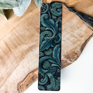 May include: A teal and black bookmark with a detailed floral design and a black tassel. The bookmark features a dark teal background with lighter teal leaves and swirls. The tassel is attached at the top.