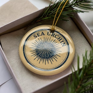 May include: A round, cream-colored ornament with the words "SEA URCHIN" in a navy blue circular design. The ornament features a navy blue sea urchin illustration. It is presented in a gift box with a sprig of evergreen.