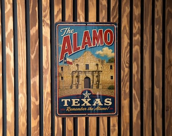 May include: A vintage-style metal sign featuring the Alamo in San Antonio, Texas. The sign has a blue background with the words "The Alamo" in red and white. The Texas flag is visible. The sign is mounted on a wooden slat wall.