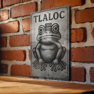 May include: A rectangular metal sign with the word "TLALOC" at the top. The sign features a detailed illustration of a frog-like creature with large eyes. The sign is gray and appears aged, mounted on a brick wall.