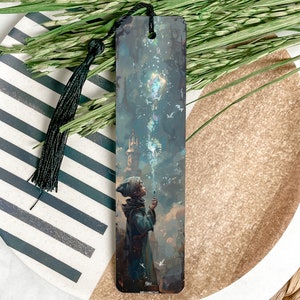 May include: A black bookmark with a colorful illustration of a person holding a glowing orb. The person is wearing a blue hooded cloak and is standing in front of a tall tower. The background is a dark blue sky with white birds flying around.