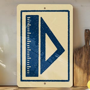May include: A rectangular sign with a cream-colored background and a dark blue illustration of a ruler and a triangle. The sign is mounted on a wooden surface, with a plant, cutting boards, and lemons in the background.
