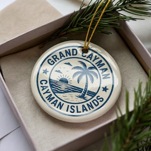 May include: A round, off-white ceramic ornament with a blue design featuring a palm tree, sun, and the text "Grand Cayman Cayman Islands." The ornament is in a gift box with a gold string for hanging and a sprig of greenery.