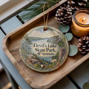 May include: A round ornament featuring a scenic landscape of Devil's Lake State Park, Wisconsin. The ornament has a gold string for hanging. The image includes a lake, cliffs, and the text "Devil's Lake State Park, Wisconsin". The ornament is on a wooden tray with pine cones and a candle.
