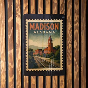 A vintage-style postage stamp from Alabama featuring a train track and a building, mounted on a wooden wall.