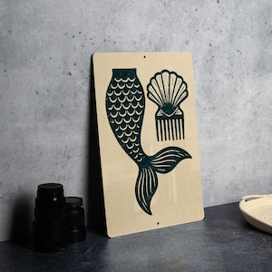 May include: A rectangular metal sign featuring a teal mermaid tail, seashell, and comb design. The artwork is printed on a beige background. The sign is leaning against a gray wall.