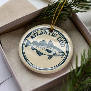 May include: A round, cream-colored Christmas ornament with a blue illustration of a fish and the words "ATLANTIC COD" in a circular border. The ornament is in a gift box with a sprig of pine.