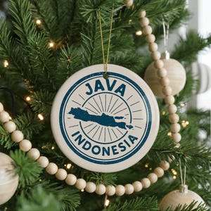 May include: A round, white ceramic Christmas ornament with a blue design featuring the words "JAVA INDONESIA" encircling a map of Java. The ornament hangs from a gold string on a Christmas tree with other ornaments and wooden bead garland.