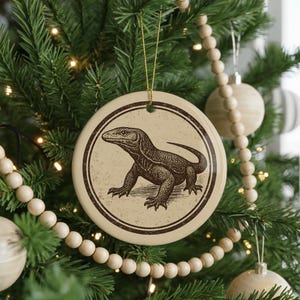 May include: A round, cream-colored Christmas ornament with a brown illustration of a Komodo dragon. The ornament is hanging on a Christmas tree with other ornaments and wooden bead garland.