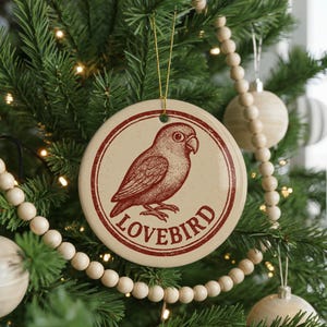 May include: A round, cream-colored Christmas ornament with a red illustration of a lovebird inside a red circular border. The word "LOVEBIRD" is printed in red below the bird. The ornament hangs from a gold string.