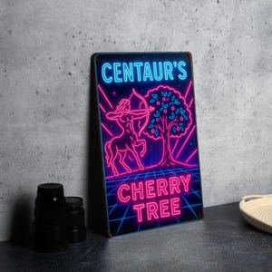 May include: A metal sign with a neon-style design. The sign features a centaur with a bow and arrow, a cherry tree, and the text "CENTAUR'S CHERRY TREE" in blue and pink. The sign has a black border and is set against a gray background.