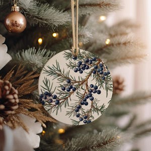 May include: A round white Christmas ornament with a sprig of juniper berries and leaves. The berries are dark blue and the leaves are green.
