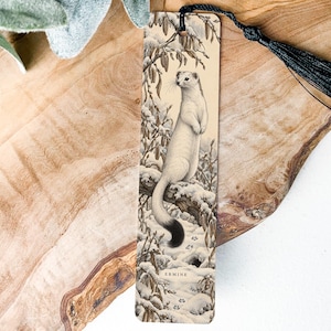 May include: A rectangular bookmark featuring a detailed illustration of a white ermine with a black-tipped tail perched on a snow-covered tree branch. The design includes winter foliage and the word ERMINE printed at the bottom. A black tassel is attached.