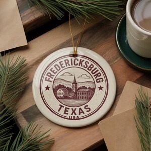 May include: A round, off-white ceramic ornament with a maroon illustration of Fredericksburg, Texas. The design includes a church, buildings, and the text "Fredericksburg Texas USA". A gold string is attached for hanging.