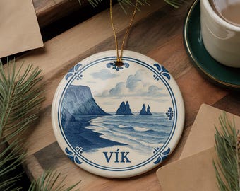 May include: A round ceramic ornament with a blue and white coastal landscape design. The ornament features a beach scene with cliffs, sea stacks, and the word "Vík". It has a gold string for hanging.