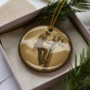 May include: A sepia-toned, round Christmas ornament featuring a vintage image of a lineman working on a utility pole. The ornament has a gold string for hanging and is presented in a gift box with evergreen sprigs.
