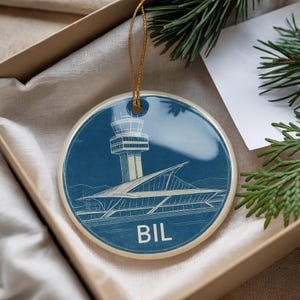 May include: Round ceramic ornament with a blue background featuring a detailed architectural illustration of an airport control tower and terminal. The ornament has a gold string for hanging and the letters "BIL" are visible.