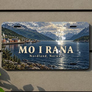 May include: A rectangular metal license plate featuring a scenic landscape of Mo i Rana, Nordland, Norway, with mountains, a body of water, and a town. The text MO I RANA and Nordland, Norway is printed in the center with four mounting holes.