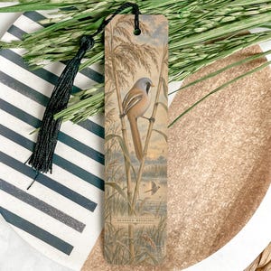 May include: A rectangular bookmark featuring a detailed illustration of a bearded reedling bird perched on tall reeds. The bookmark has a tan background, a hole at the top with a black tassel attached, and the text "BEARDED REEDLING" printed at the bottom.