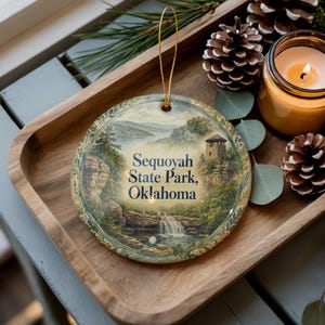 May include: Round ceramic ornament featuring a scenic illustration of Sequoyah State Park, Oklahoma. The image includes a waterfall, tower, and lake. The ornament has a gold string for hanging and is displayed on a wooden tray with pinecones and a lit candle.