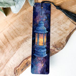 May include: A rectangular bookmark featuring a detailed illustration of a blue lantern with a warm, glowing interior. The background is a dark, starry night sky. A black tassel hangs from the top. The bookmark is placed on a wooden surface.