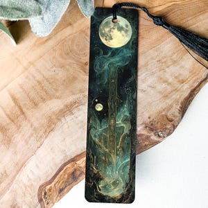 May include: A black bookmark with a green and gold illustration of a moon, stars, and a tree. The bookmark has a black tassel.