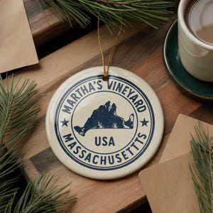 May include: A round, off-white ceramic ornament with a gold string for hanging. The ornament features a blue illustration of Martha's Vineyard, Massachusetts, with the text "MARTHA'S VINEYARD" and "MASSACHUSETTS USA".