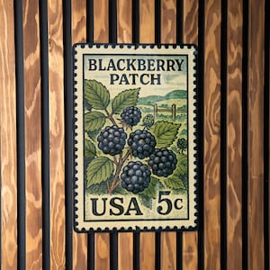 May include: A vintage-style sign featuring a blackberry patch design, resembling a postage stamp. The sign displays the text "Blackberry Patch" and "USA 5c" with illustrations of blackberries and leaves. The sign is mounted on a wooden slat wall.