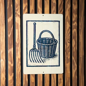 May include: A rectangular sign with a blue illustration of a rake and a basket. The sign is mounted on a wooden wall with vertical brown and black planks. The illustration has a vintage, woodcut style.