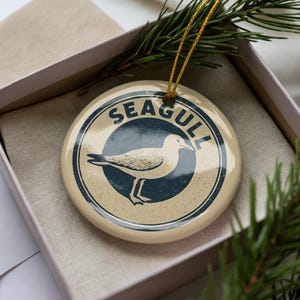 May include: A round, cream-colored ornament with a seagull illustration inside a navy blue circle. The word "SEAGULL" is printed in navy blue around the circle. The ornament is in a gift box with a sprig of pine.