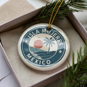 May include: A round, white ceramic ornament featuring a beach scene with a palm tree, sun, and the text "Isla Mujeres Mexico" in a blue and white design. The ornament is in a gift box with a gold string.