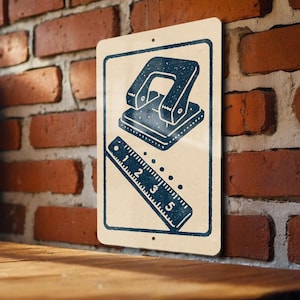 May include: A rectangular sign with a cream background and a dark blue illustration of a hole punch and a ruler. The sign is mounted on a red brick wall and a wooden surface.