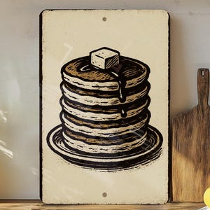 May include: A vintage-style metal sign featuring a stack of pancakes with syrup and a pat of butter. The illustration is in shades of brown and black, set against a cream-colored background. The sign has a rustic, aged appearance.