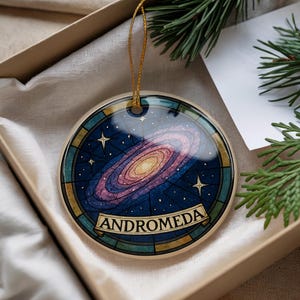 May include: Round Christmas ornament featuring a colorful depiction of the Andromeda galaxy. The ornament has a gold string for hanging and is presented in a gift box. The design includes stars and the word "ANDROMEDA".