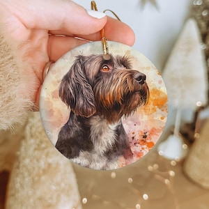 May include: A round ceramic ornament featuring a watercolor painting of a brown and black dog with a white chest. The dog is looking to the right and has a soft, fuzzy appearance. The background is a light orange and pink watercolor wash.
