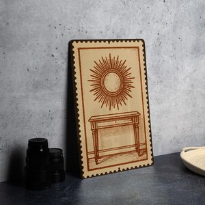 May include: A rectangular wooden sign with a brown border and a beige background. The sign features a brown illustration of a sunburst above a table with a drawer. The sign is leaning against a gray wall.