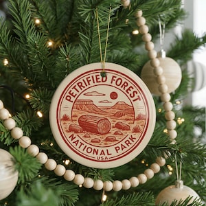 May include: A round, cream-colored Christmas ornament with a red illustration of Petrified Forest National Park. The design includes a landscape with mountains, a log, and the text "PETRIFIED FOREST NATIONAL PARK USA". The ornament hangs from a gold string.