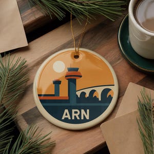 May include: Round ceramic ornament with a retro-style illustration of a control tower, bridge, and sun in shades of orange, blue, and beige. The letters "ARN" are at the bottom. A gold string is attached for hanging.