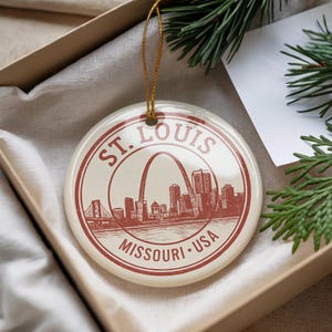May include: A round ceramic Christmas ornament featuring a red and white design of the St. Louis skyline, including the Gateway Arch. The ornament is suspended by a gold string and is presented in a gift box.