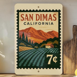 May include: A rectangular sign with a vintage postage stamp design featuring the text "San Dimas California" and a landscape illustration of mountains, fields, and a house. The sign has a cream-colored background and a dark green border.