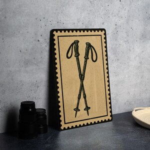 May include: A framed print featuring a vintage-style illustration of two crossed hiking poles. The artwork is set against a tan background with a black border, resembling a postage stamp. The poles are dark green, and the overall aesthetic is rustic.
